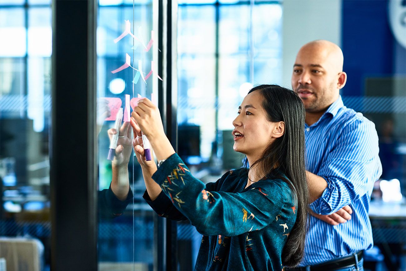 Asian woman hanging sticky note while man standing behind her watches